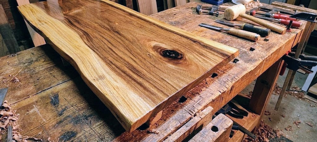 Wooden workbench with a large wooden slab and various tools on a workshop floor.